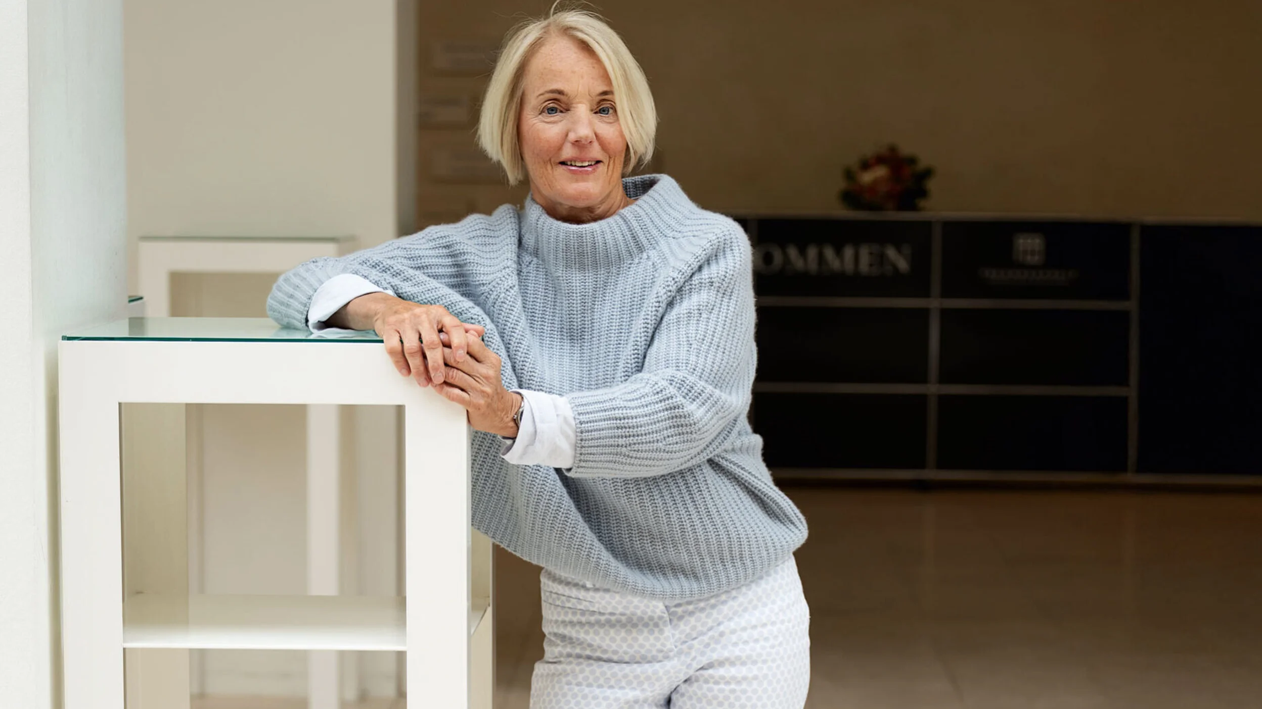 Eine Person lehnt sich leger an einen hohen Tisch, trägt weiße Hose und grauen Pullover, in einem modernen, hellen Raum.