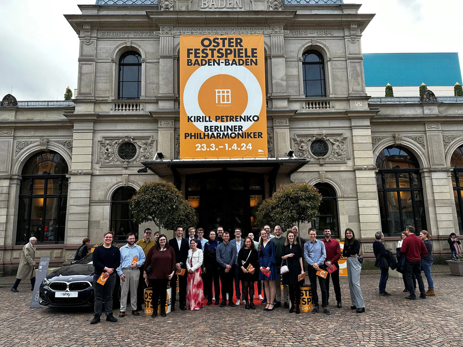 Eine Gruppe von Menschen steht vor dem Festspielhaus mit einem großen Plakat für die Osterfestspiele 2024.