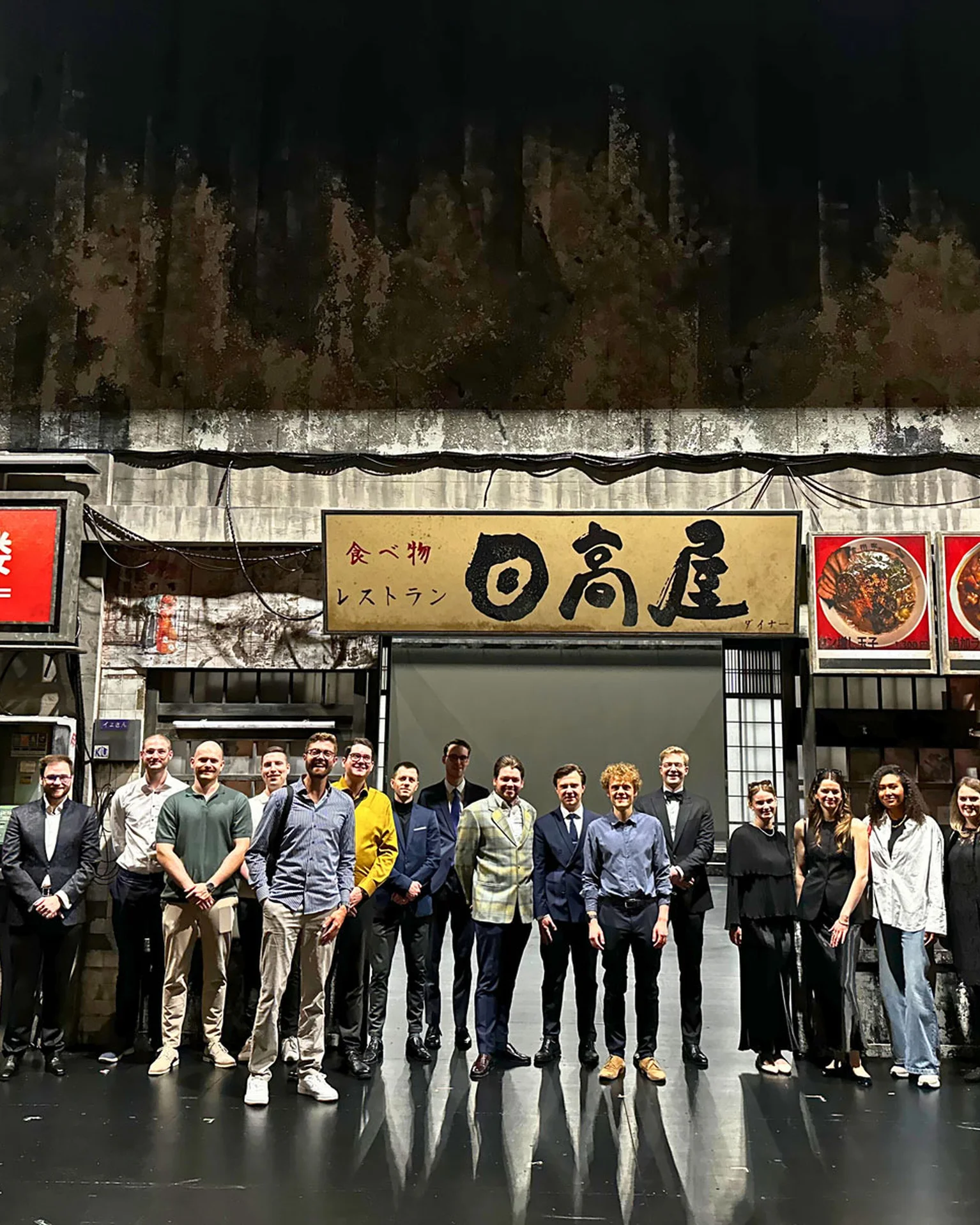 Eine Gruppe von Menschen posiert auf einer Bühne vor der Kulisse zu "Madama Butterfly", die eine asiatische Straßenszene darstellen. Verschiedene Schilder sind sichtbar.