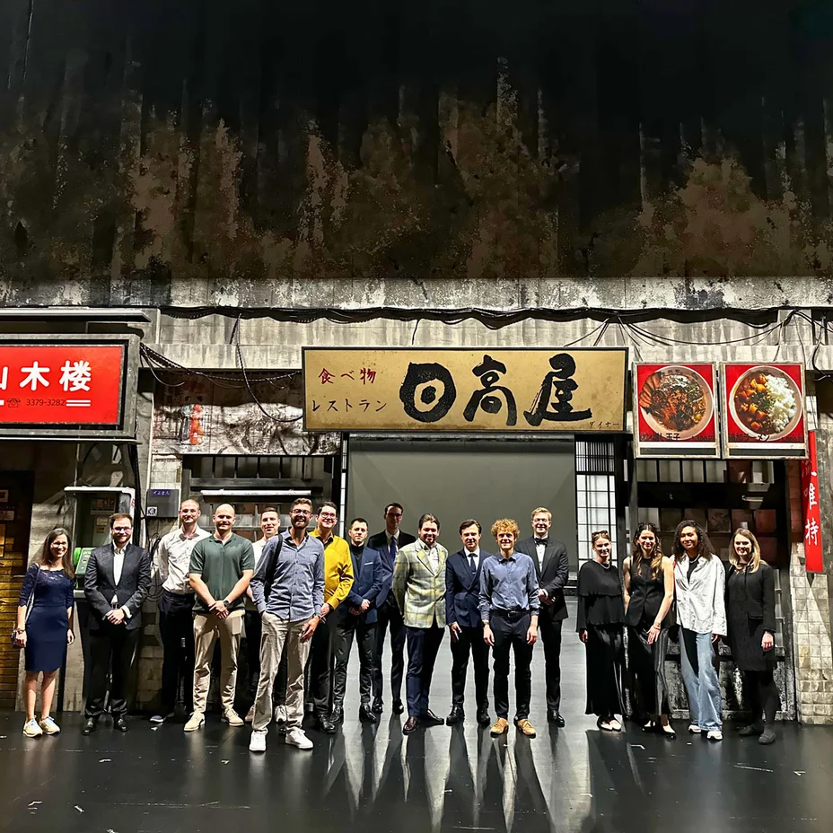 Eine Gruppe von Menschen posiert auf einer Bühne vor der Kulisse zu "Madama Butterfly", die eine asiatische Straßenszene darstellen. Verschiedene Schilder sind sichtbar.