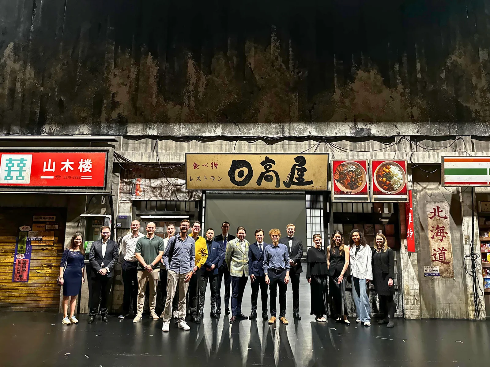 Eine Gruppe von Menschen posiert auf einer Bühne vor der Kulisse zu "Madama Butterfly", die eine asiatische Straßenszene darstellen. Verschiedene Schilder sind sichtbar.