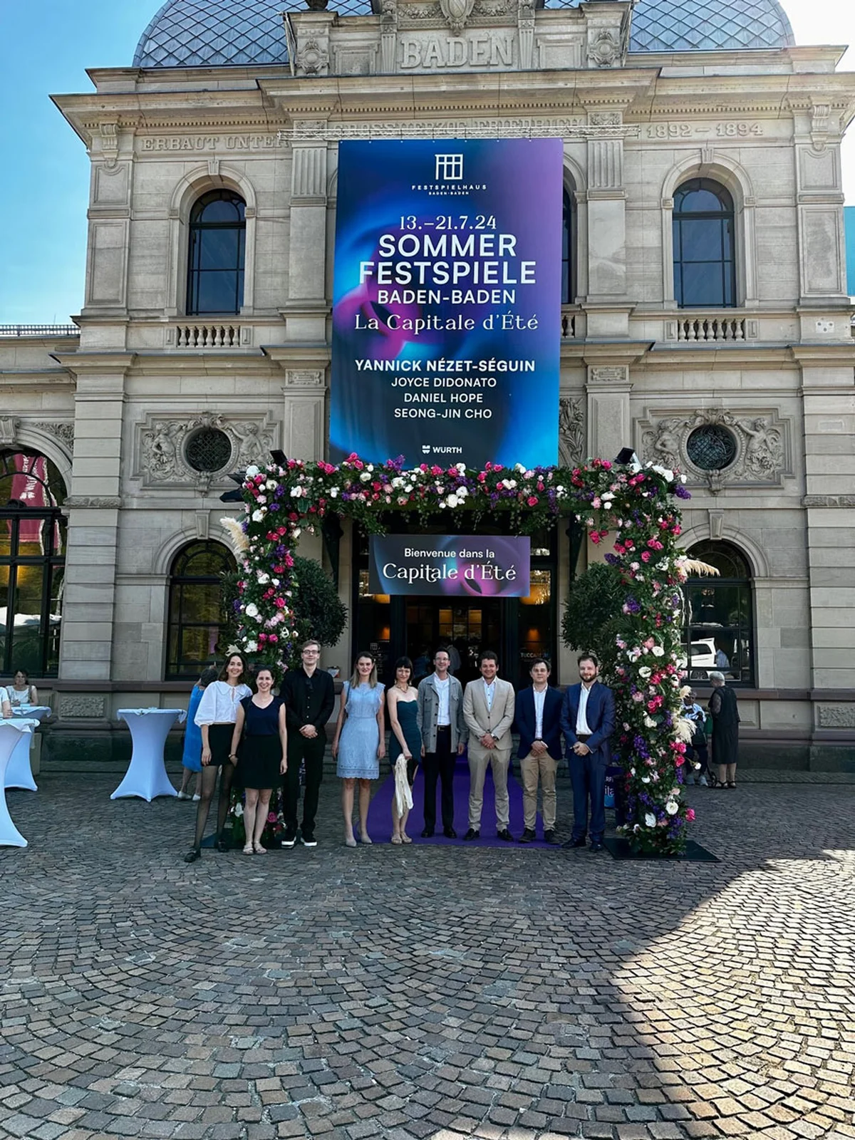 Teilnehmer stehen vor dem Festspielhaus mit großem Banner der Sommerfestspiele Baden-Baden, umgeben von Blumenarrangements.