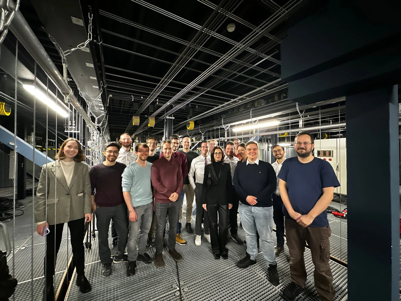 Eine Gruppe von Menschen steht in einem industriellen Raum mit sichtbaren Leitungen und Metallstrukturen im Hintergrund. Alle blicken in die Kamera.