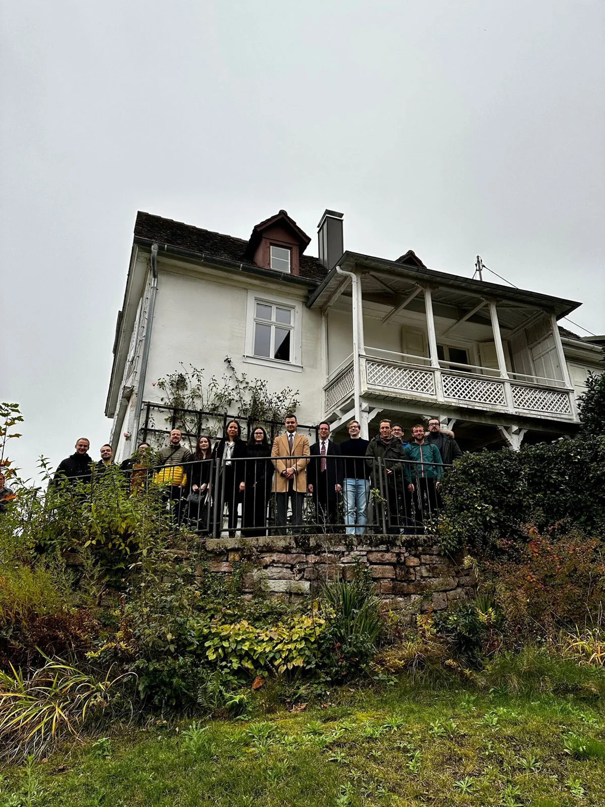 Eine Gruppe von Menschen steht auf einer Terrasse vor dem weißen Brahms-Haus, umgeben von üppigem Grün und bewölktem Himmel.