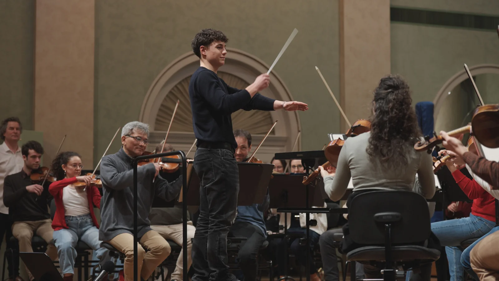 Ein Schüler dirigiert ein Orchester, während Musiker mit Streichinstrumenten spielen. Der Raum ist elegant mit klassischer Architektur.