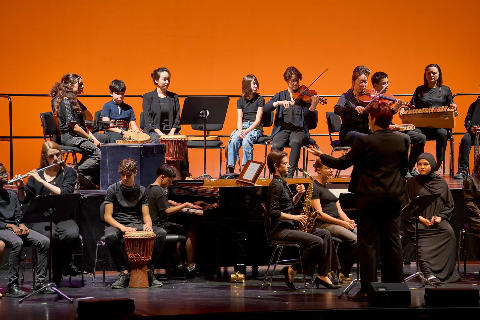 Ein Orchester aus Schülern spielt auf einer Bühne vor einem orangefarbenen Hintergrund. Musiker verwenden verschiedene Instrumente, während der Dirigent leitet.