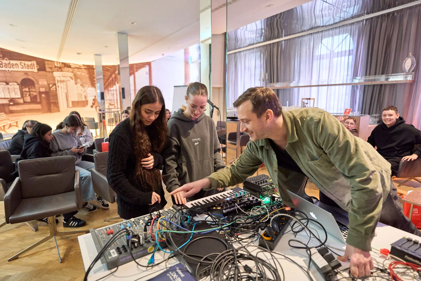 Personen bei einem Workshop mit Audiomischpulten und Kabeln. Ein Mann erklärt etwas, umgeben von jungen Teilnehmern in einem modernen Raum.