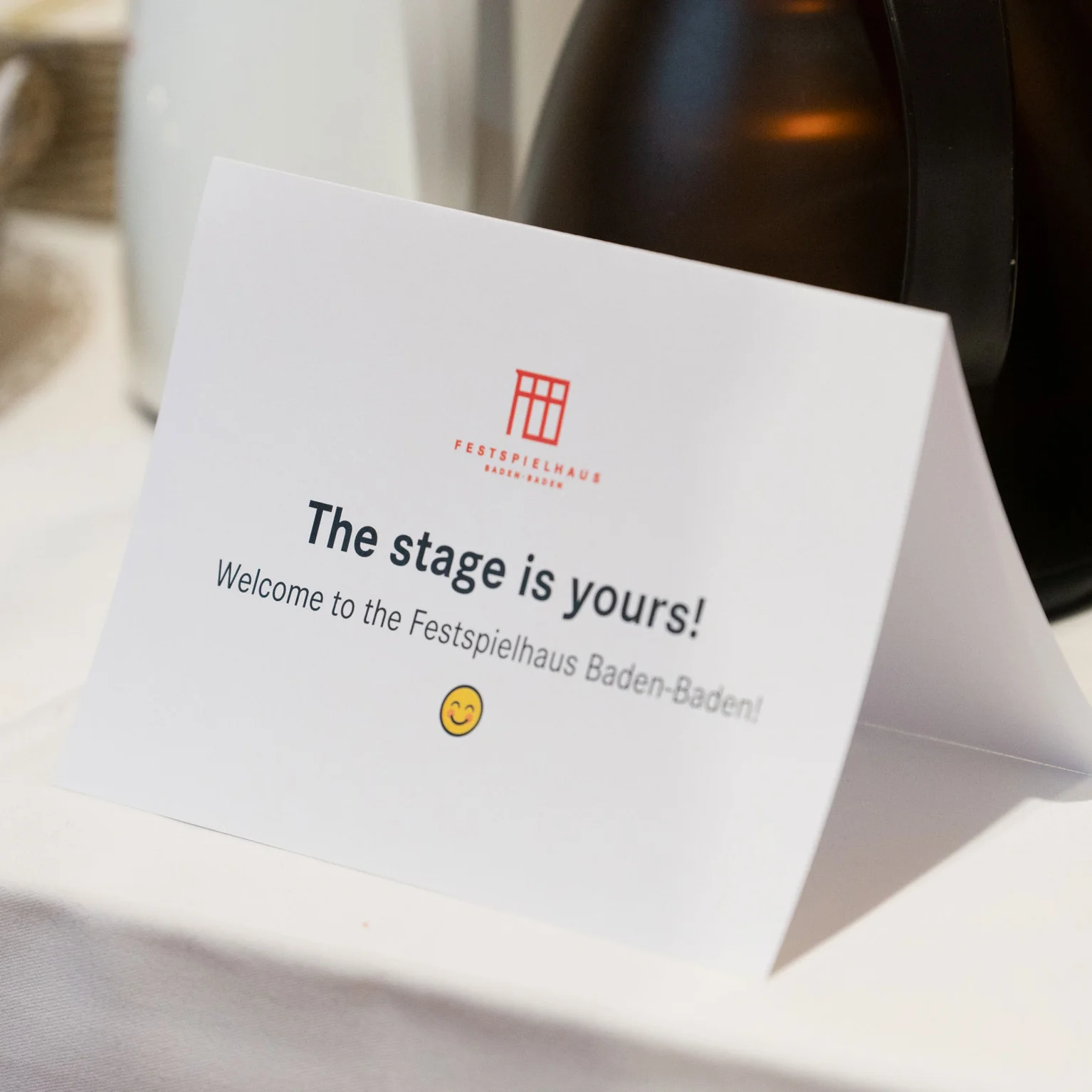 Ein Tischschild mit der Aufschrift „The stage is yours! Welcome to the Festspielhaus Baden-Baden“, neben Getränkeflaschen.