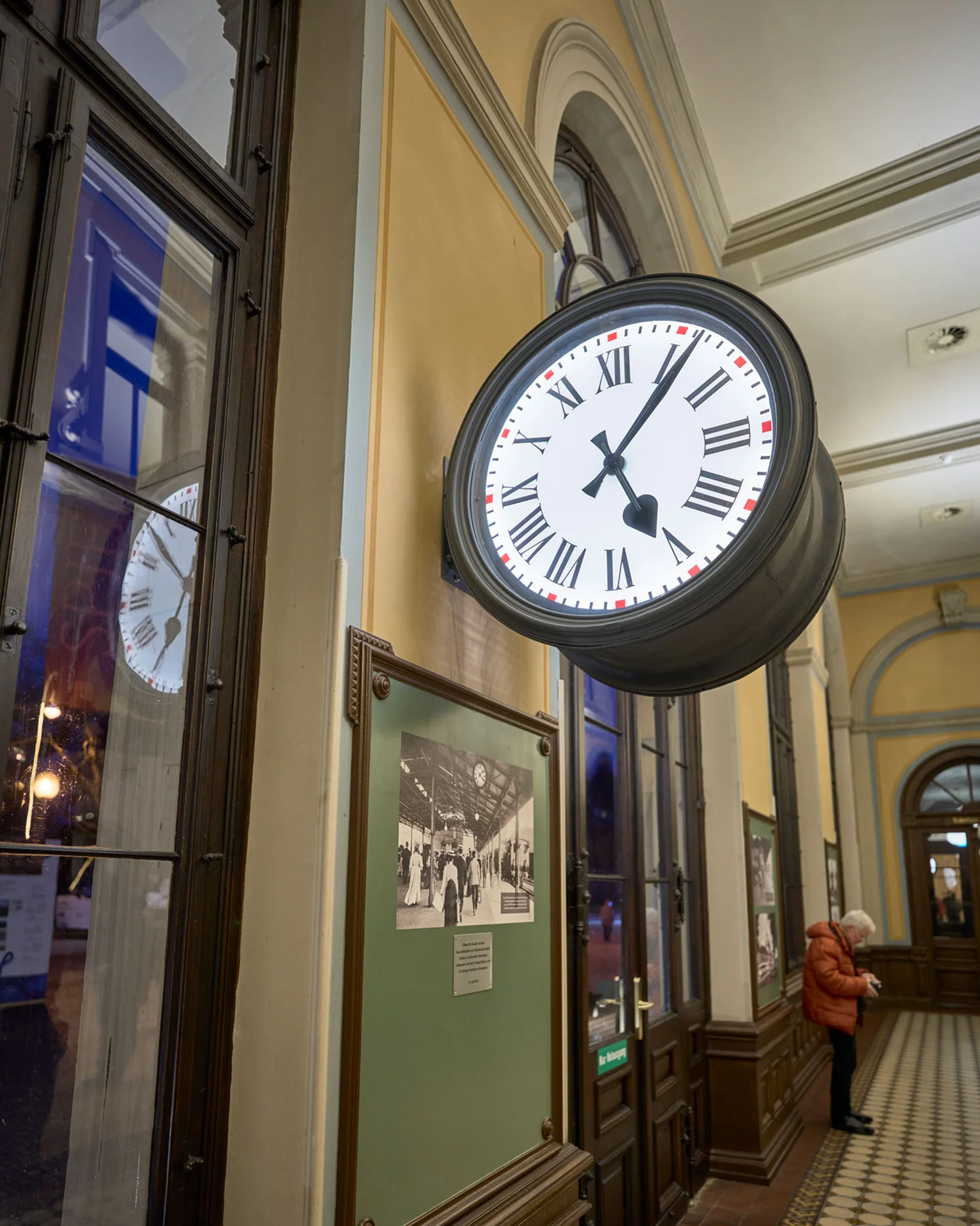 Ein großer Bahnhofsuhr hängt an der Wand in einem historischen Gebäude mit korinthischen Säulen.