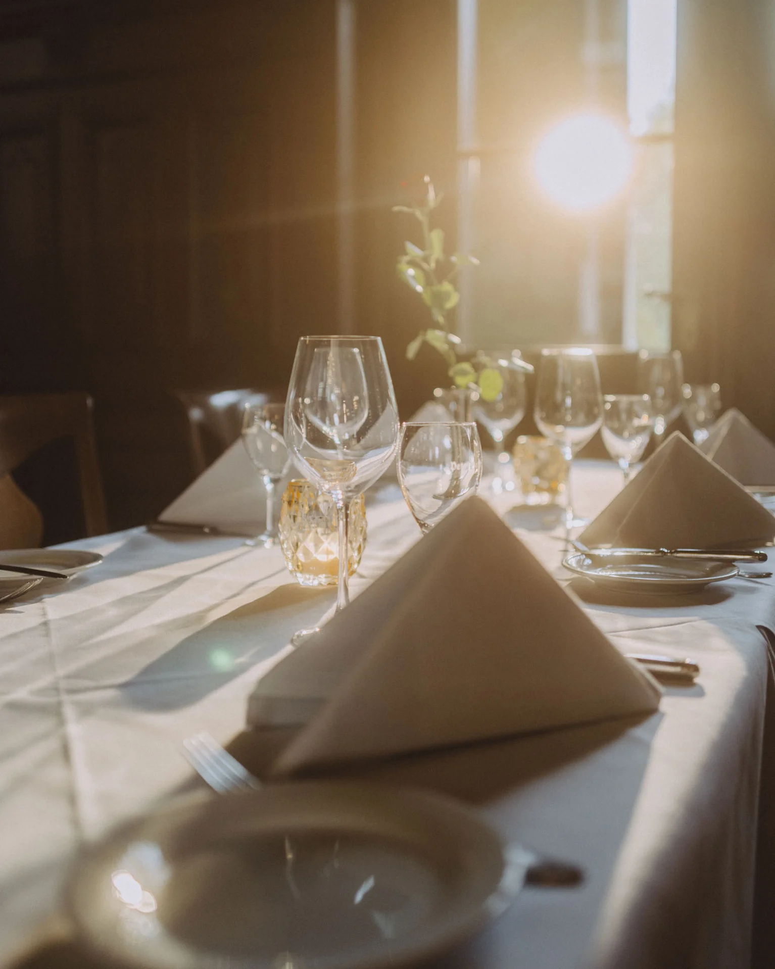 Ein stilvoll gedeckter Tisch mit Gläsern und Stoffservietten, sanft beleuchtet von warmem Sonnenlicht, das durch ein Fenster scheint.
