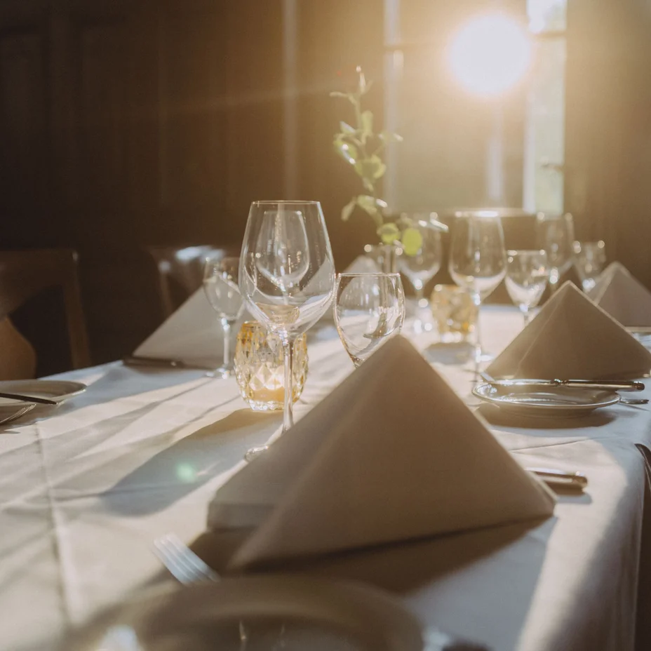 Ein stilvoll gedeckter Tisch mit Gläsern und Stoffservietten, sanft beleuchtet von warmem Sonnenlicht, das durch ein Fenster scheint.