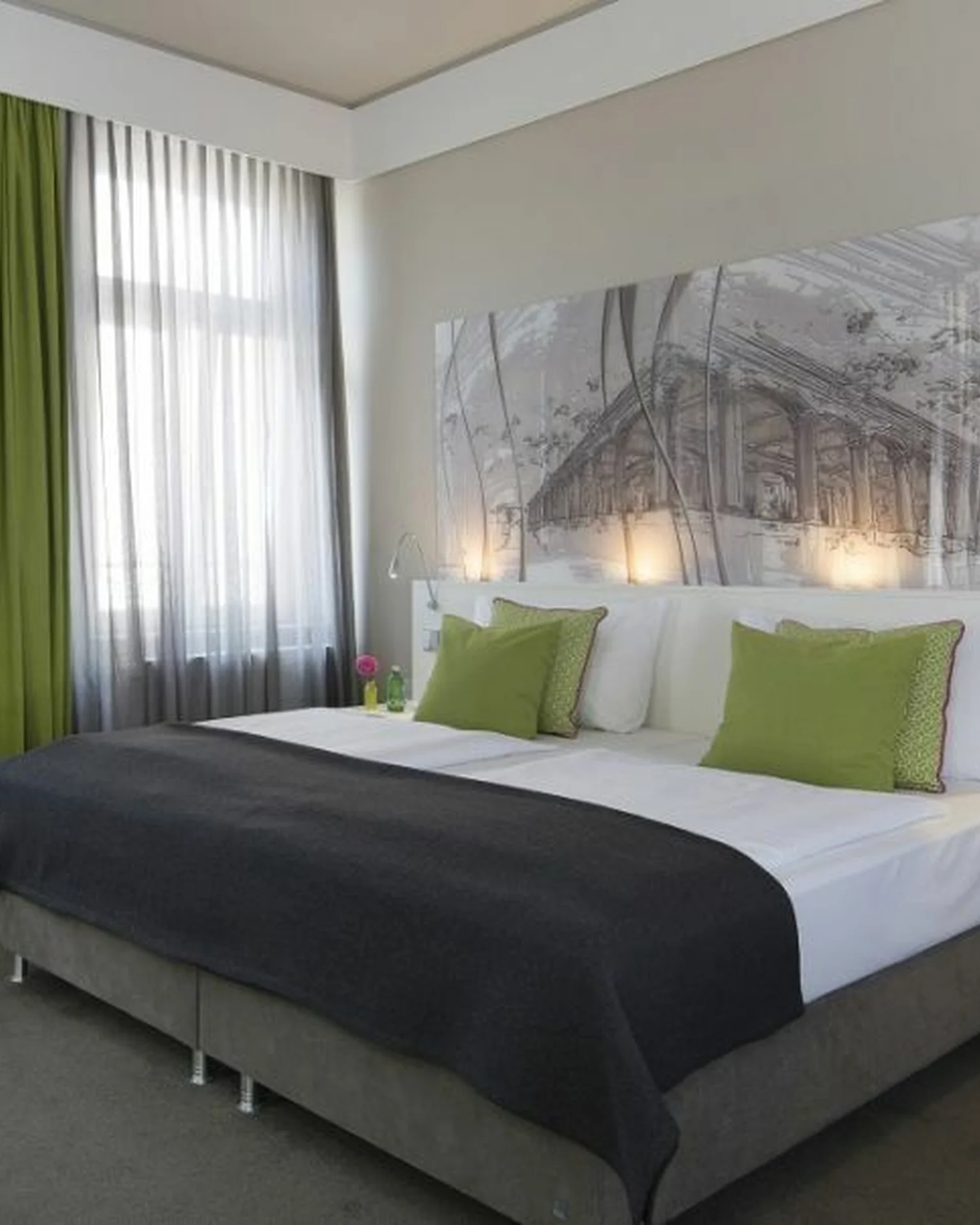 Modernes Hotelzimmer mit grauem Bett und grünen Kissen. Weiße Vorhänge und dekorative Wandbeleuchtung sorgen für eine einladende Atmosphäre.