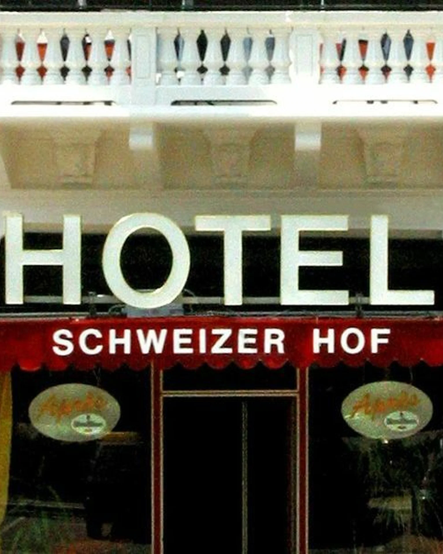 Eingang des Hotels Schweizer Hof mit rotem Vordach und weißen Balkonen. Die Fassade ist in Rot und Weiß gehalten.