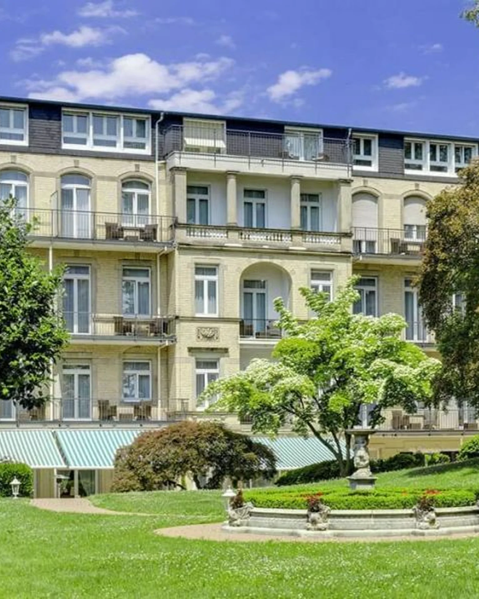Elegantes, historisches Gebäude aus mehreren Etagen mit Giebeldach, umgeben von gepflegter Gartenlandschaft, Bäumen und Sitzbänken unter klarem blauem Himmel.