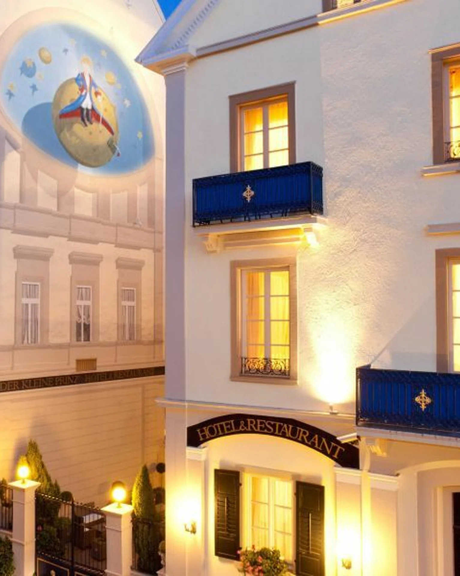 Abendansicht einer belebten Straße mit eleganten, beleuchteten Gebäuden. Ein großes Wandgemälde und dekorative Balkone sind zu sehen.