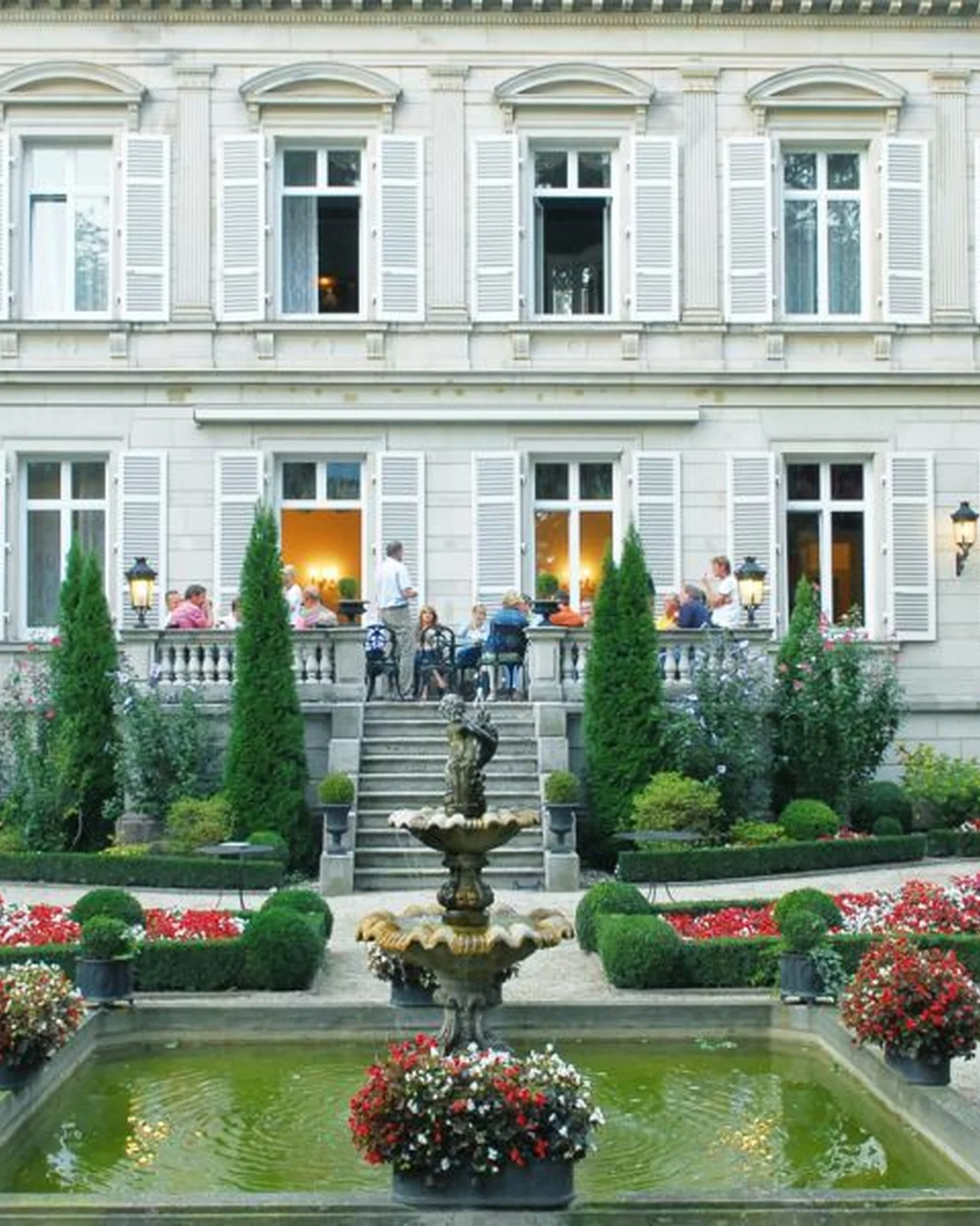 Prächtiges historisches Gebäude mit gepflegtem Garten und Brunnen. Menschen stehen auf der Treppe vor dem Eingang und genießen die Atmosphäre.