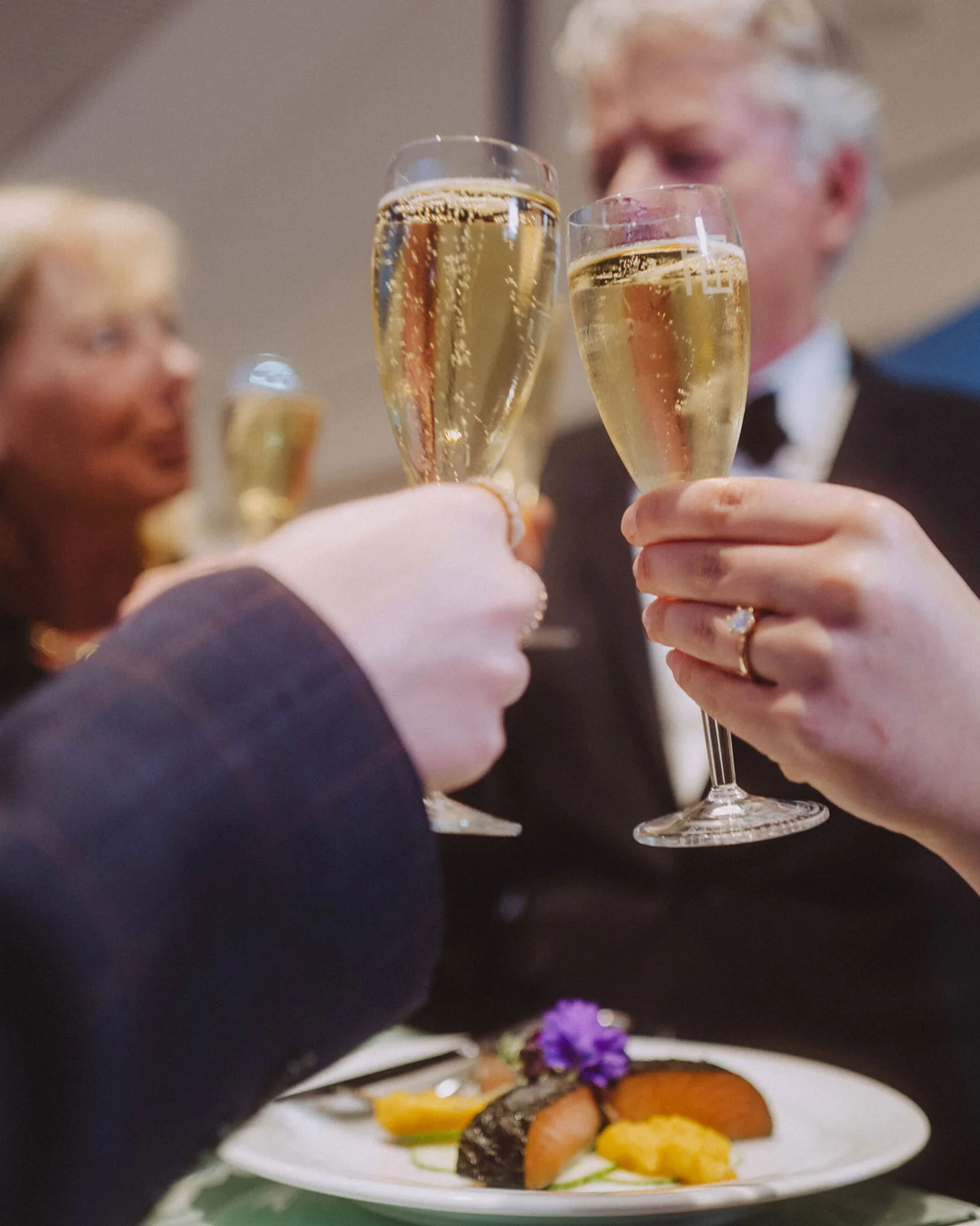 Gruppe von Menschen stößt mit gefüllten Champagnergläsern an. Im Vordergrund steht ein Teller mit dekorativ angerichteten Speisen.