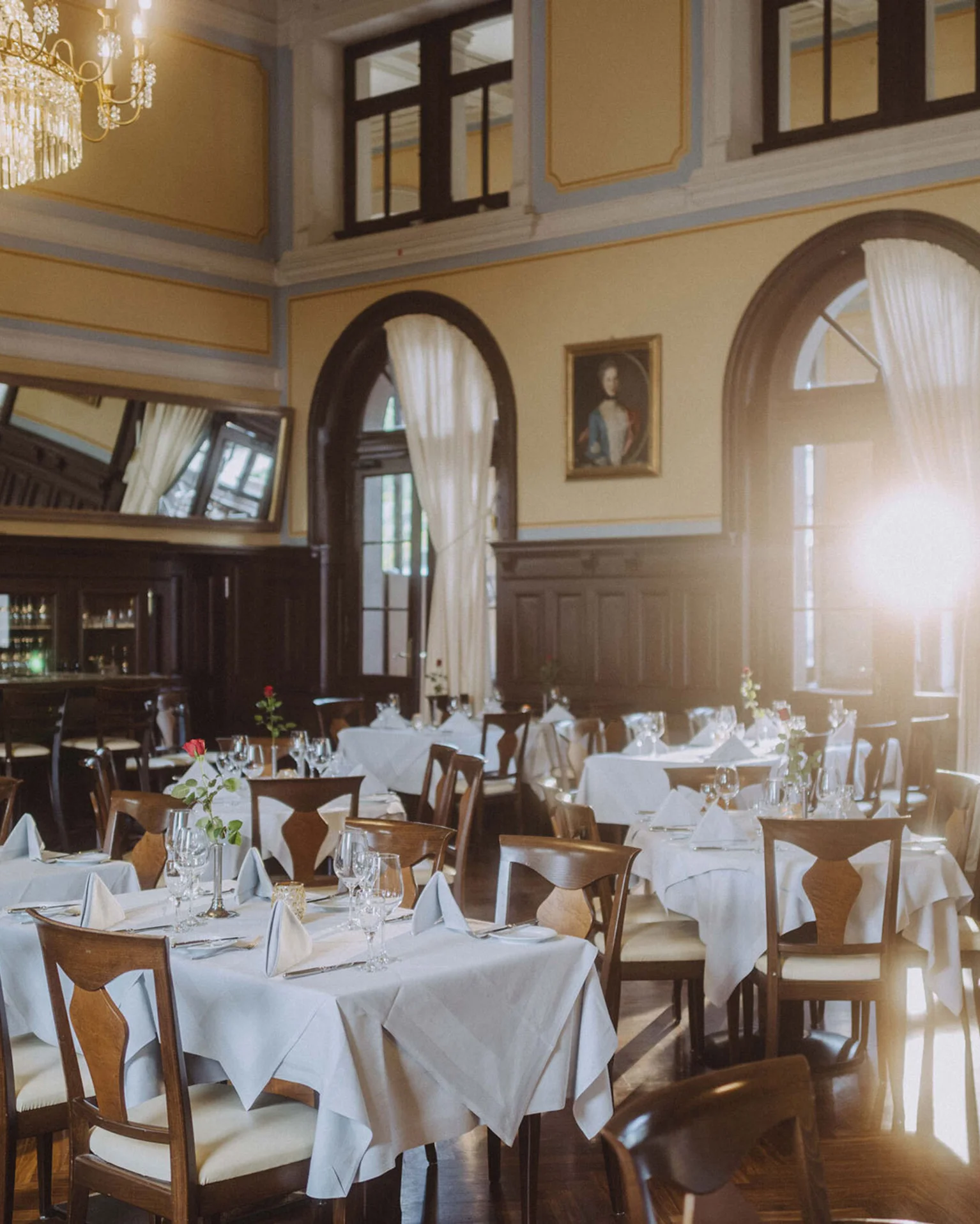 Eleganter Speisesaal mit gedeckten Tischen, Kronleuchter und gewölbten Fenstern. Die Sonne strahlt durch Glas, blumige Tischdekoration.