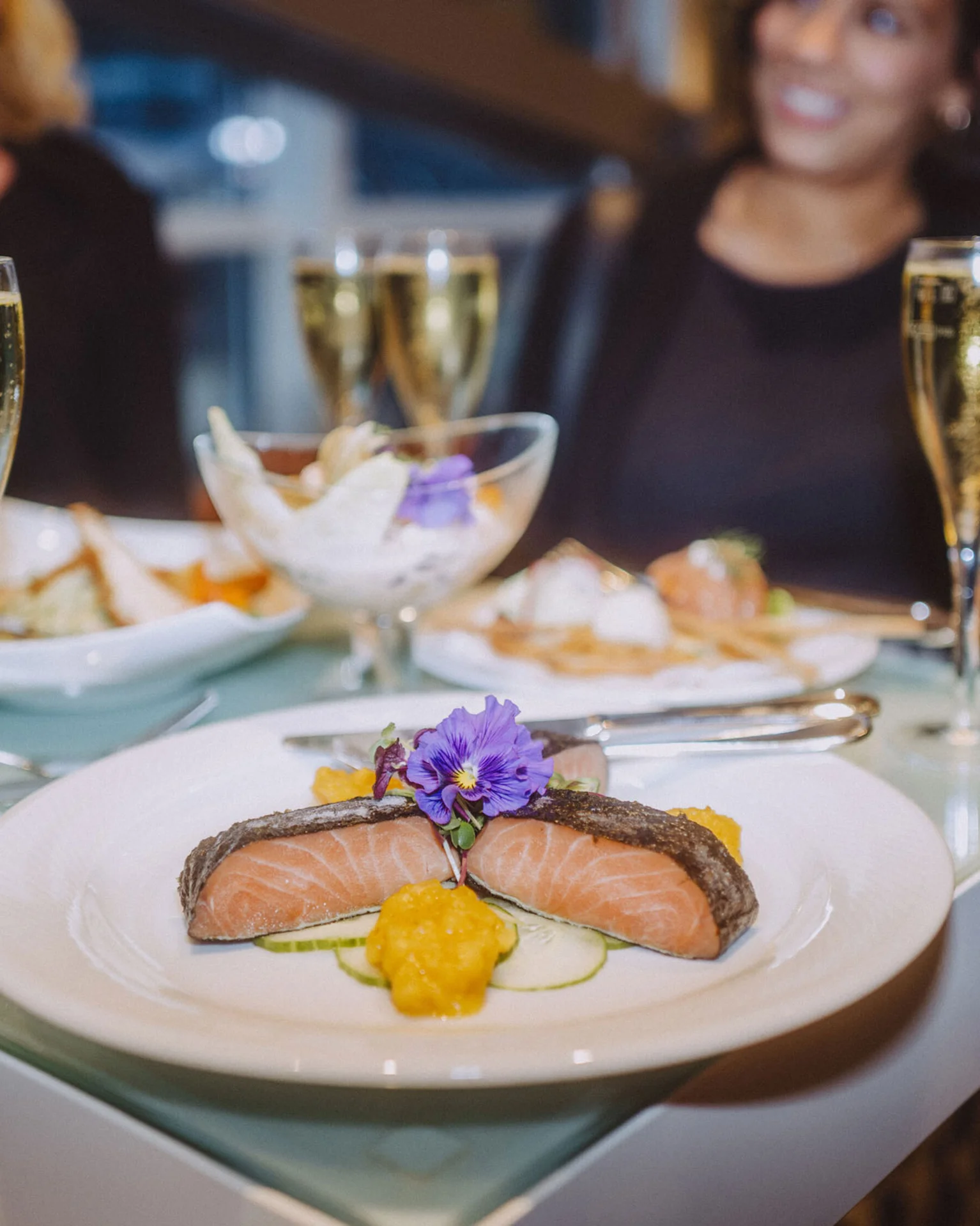 Ein eleganter Tisch mit einem Gourmetgericht aus Lachs und Gemüse, dekoriert mit Blüten, umgeben von Sektgläsern.