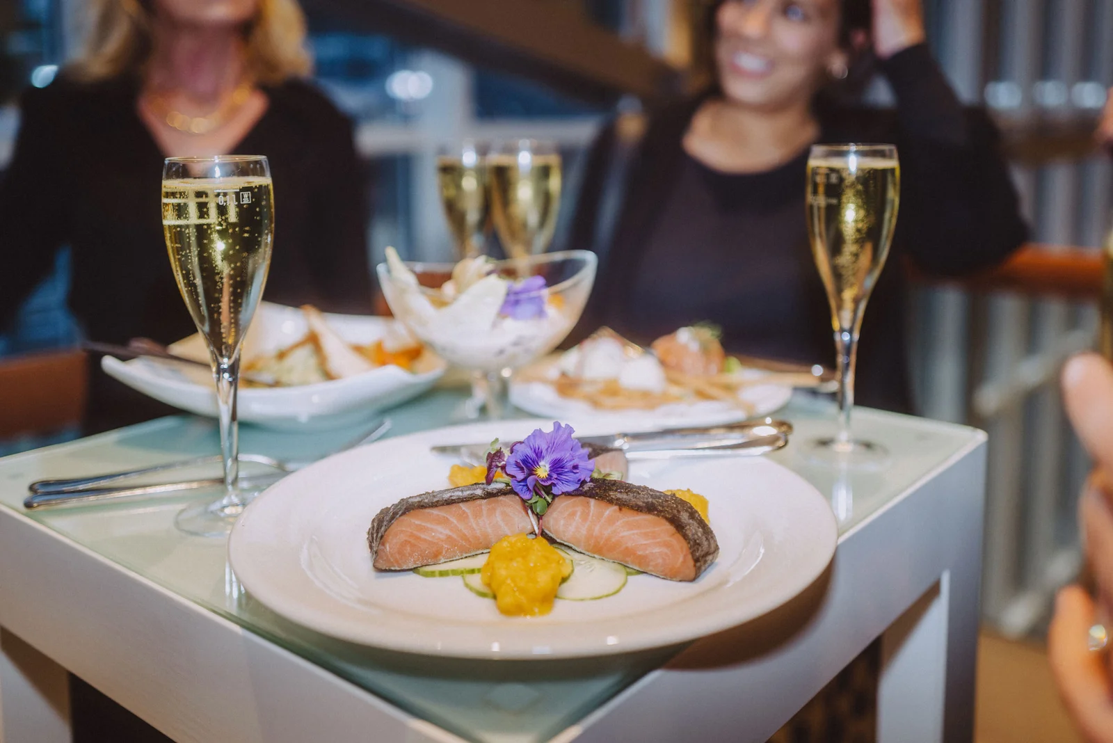Ein eleganter Tisch mit einem Gourmetgericht aus Lachs und Gemüse, dekoriert mit Blüten, umgeben von Sektgläsern.