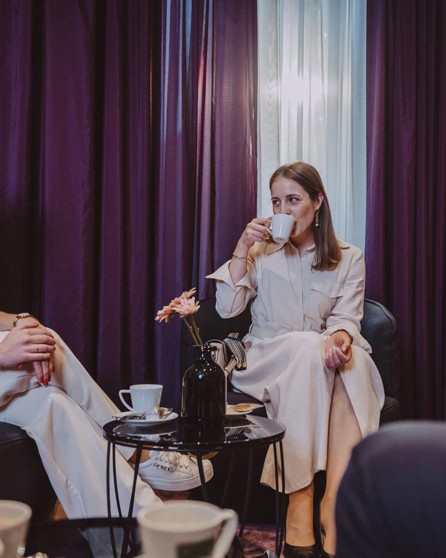 Drei Personen sitzen in einem gemütlichen Raum mit lila Vorhängen, trinken Kaffee und unterhalten sich entspannt.
