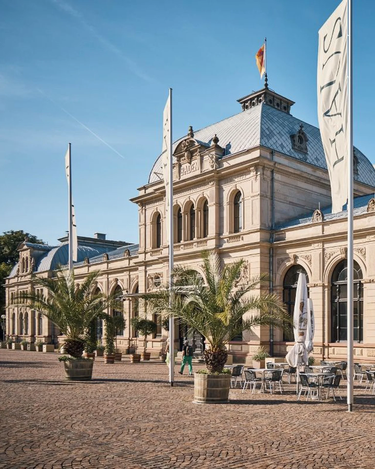 Historisches Gebäude mit Sandsteinfassade und hohen Fenstern. Vor dem Eingangsbereich stehen Fahnen und Topfpflanzen. Ruhige und sonnige Atmosphäre.
