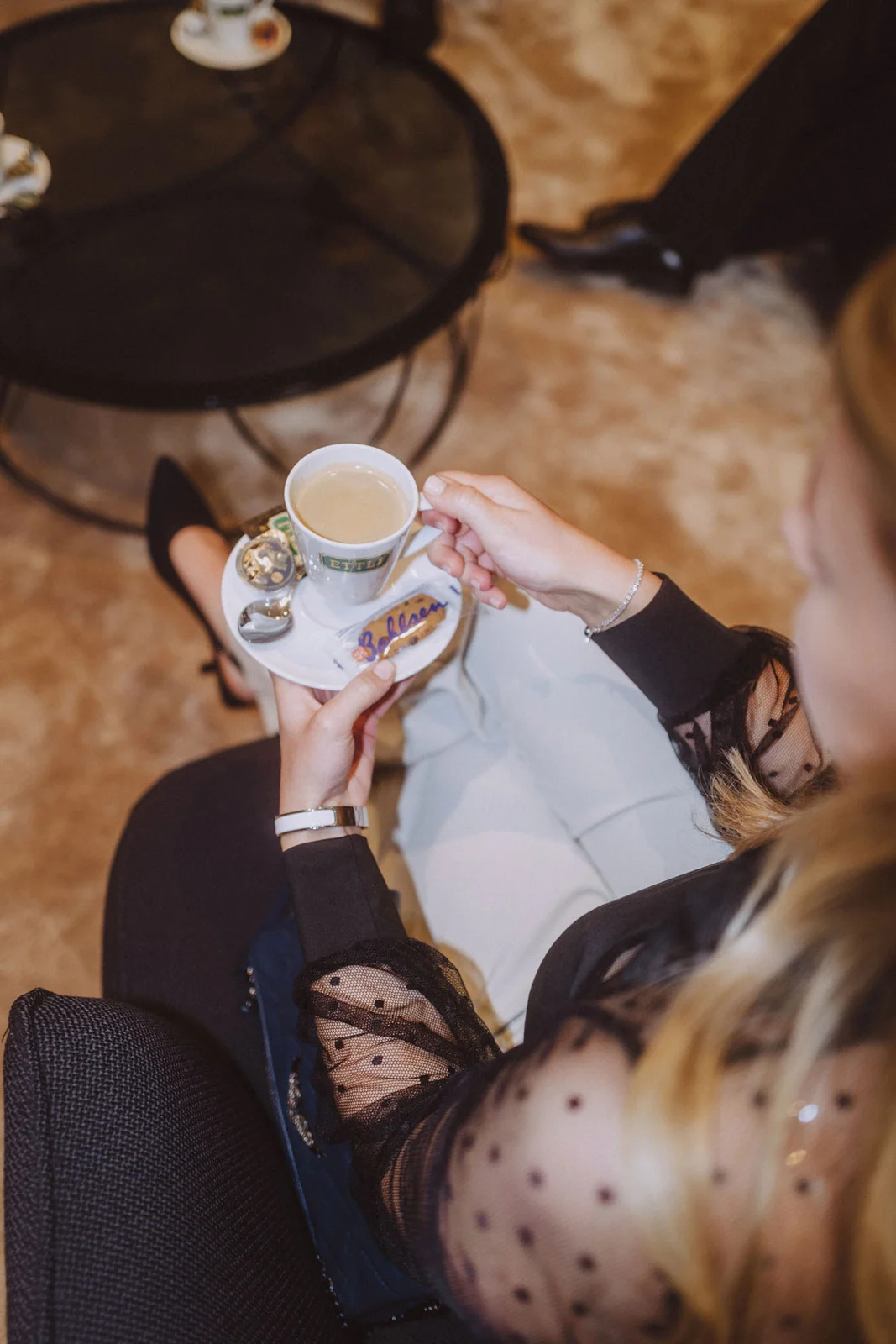 Eine Person hält eine Tasse Kaffee auf einer Untertasse mit einem Stück Gebäck. Elegantes Ambiente mit einem Teppichboden.