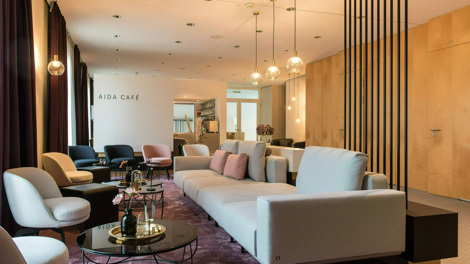 Ein eleganter Loungeraum mit modernen Sofas und Sesseln auf einem lila Teppich. Dezente Beleuchtung und helle Holzakzente dominieren das Design.