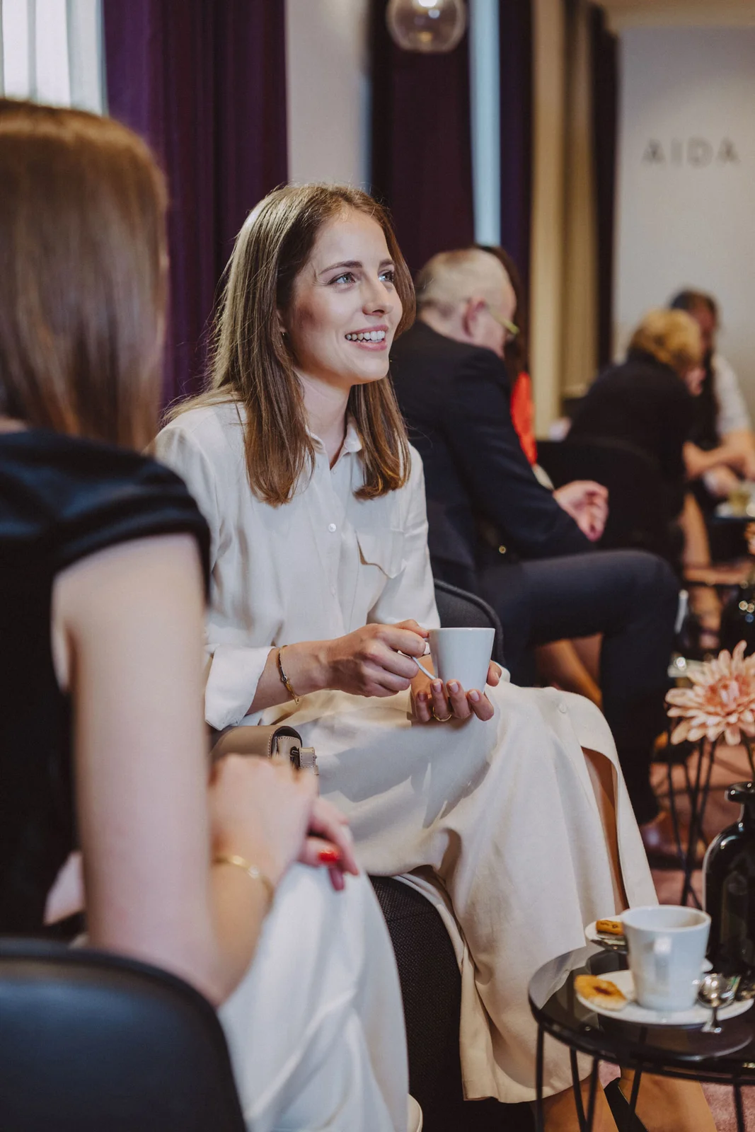 Person in weißem Kleid hält eine Tasse, während sie mit anderen auf einem Event in einem eleganten Raum spricht.