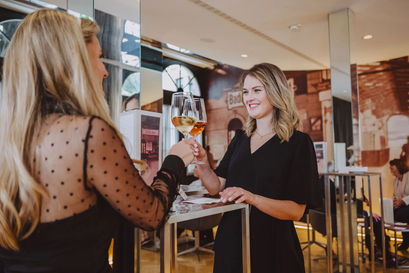 Zwei Frauen stoßen mit Sektgläsern in einem elegant beleuchteten Raum an. Beide tragen schwarze Kleider.