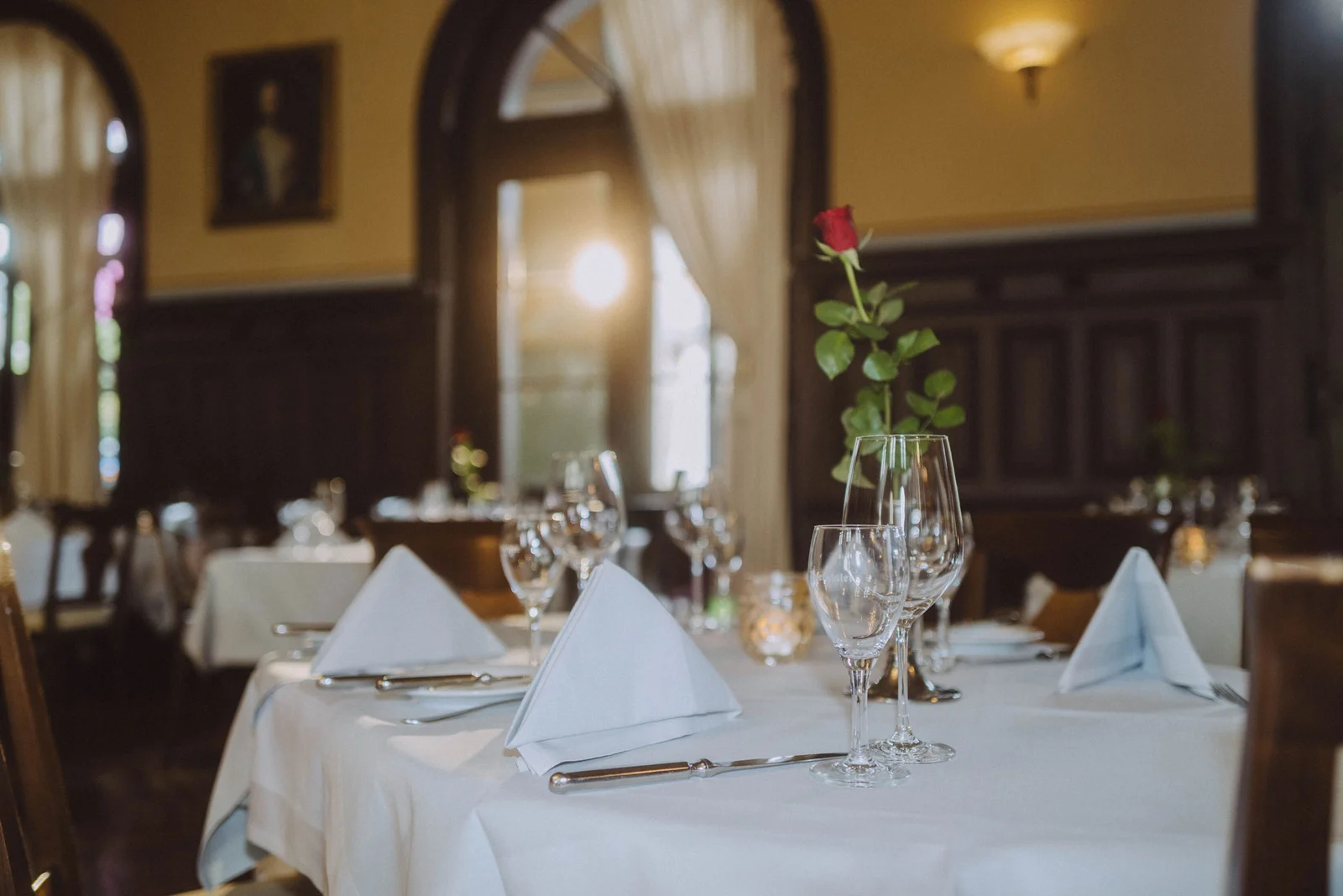 Elegantes Restaurant mit gedecktem Tisch, weißen Servietten und einer roten Rose in einem Glasgefäß. Weiche Beleuchtung und Vorhänge im Hintergrund.