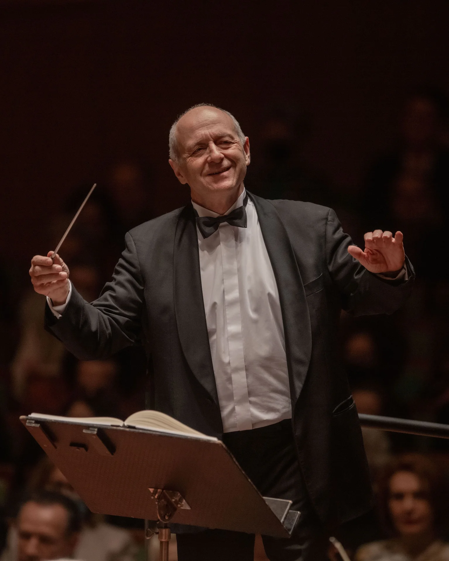 Ein Dirigent im formellen Anzug dirigiert ein Orchester. Er hält den Taktstock und ist umgeben von konzentrierten Musikern.