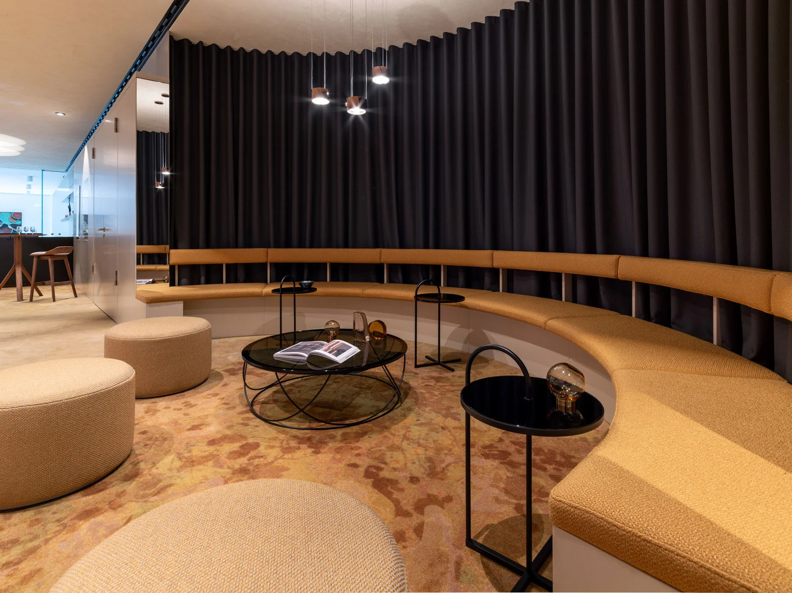 Moderner Loungebereich mit geschwungenem, cremefarbenem Sofa, runden Tischen und grauen Vorhängen. Dezente Beleuchtung und beige Teppichboden.