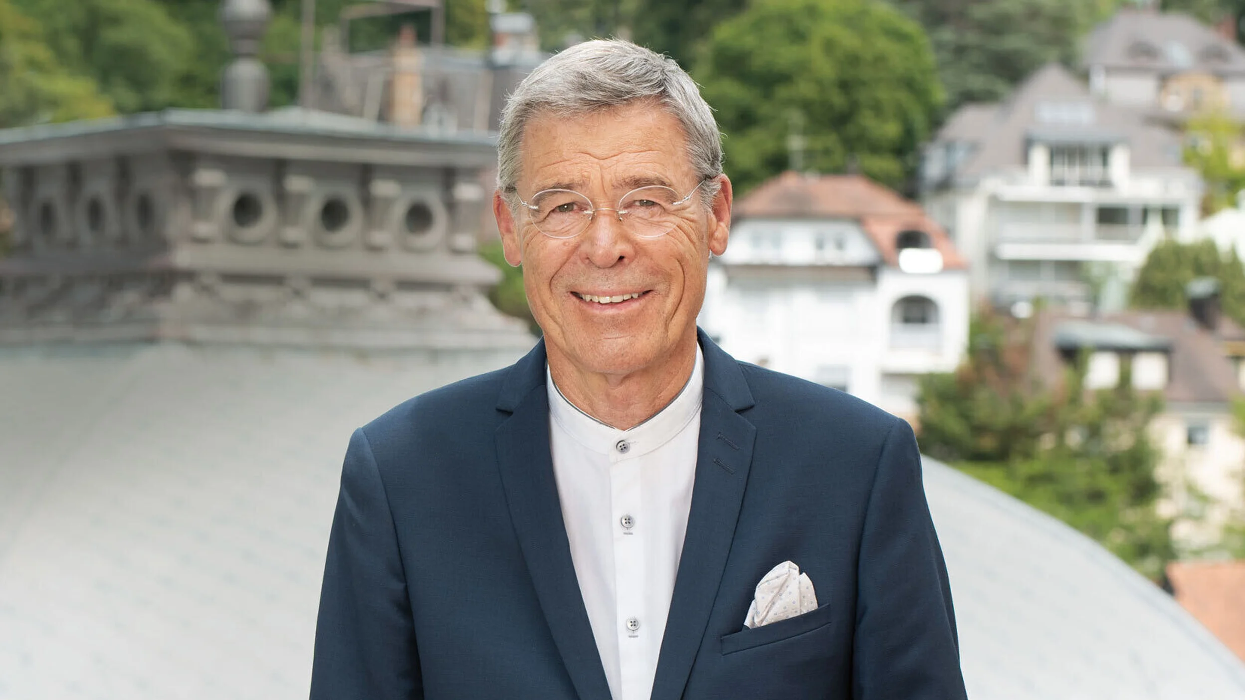 Ein älterer Mann steht in einem eleganten Anzug auf einem Balkon mit Blick auf eine städtische Landschaft im Hintergrund.