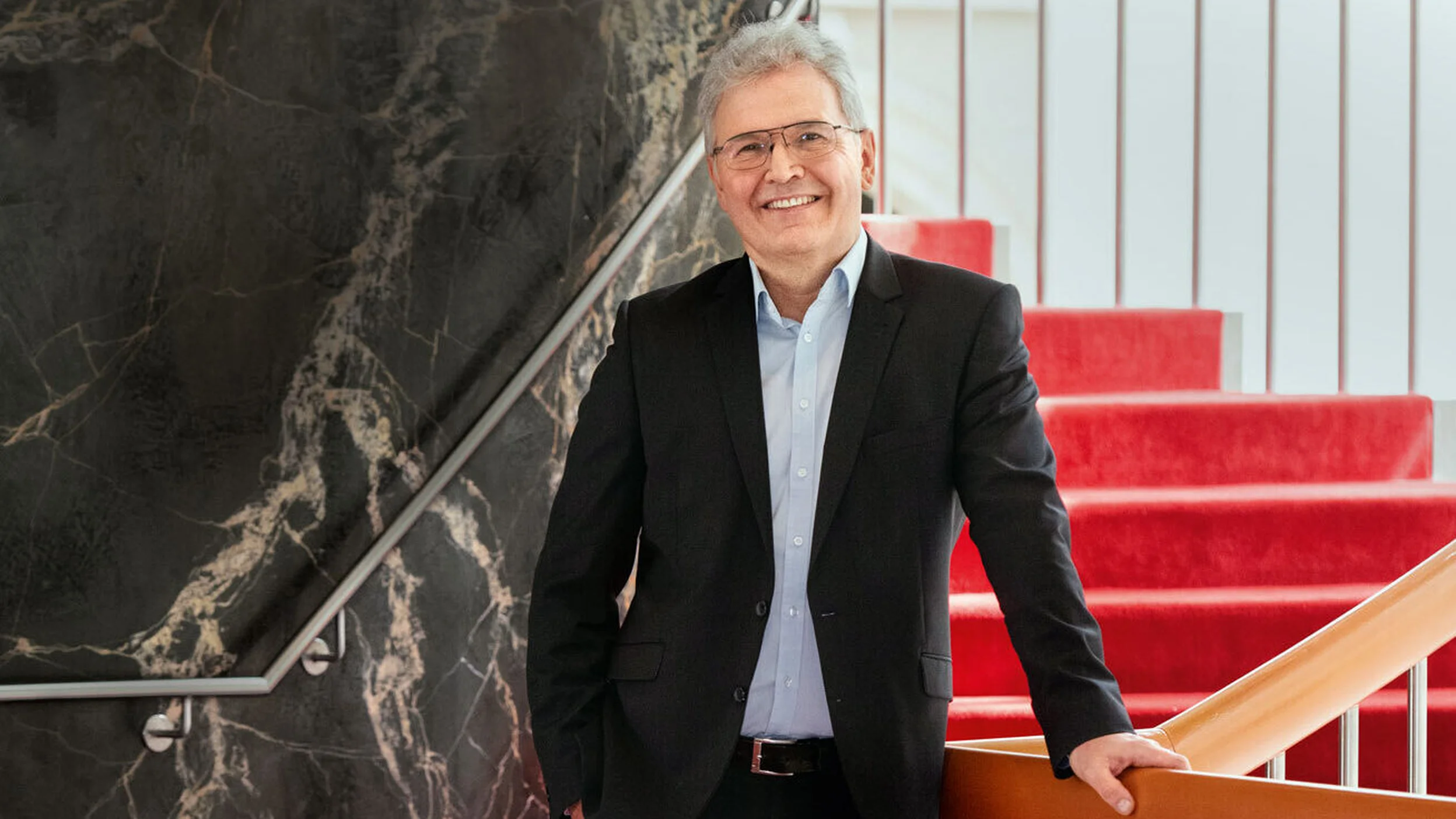 Ein Mann im schwarzen Anzug steht auf einer roten Treppe in einem modernen Gebäude und lächelt freundlich.
