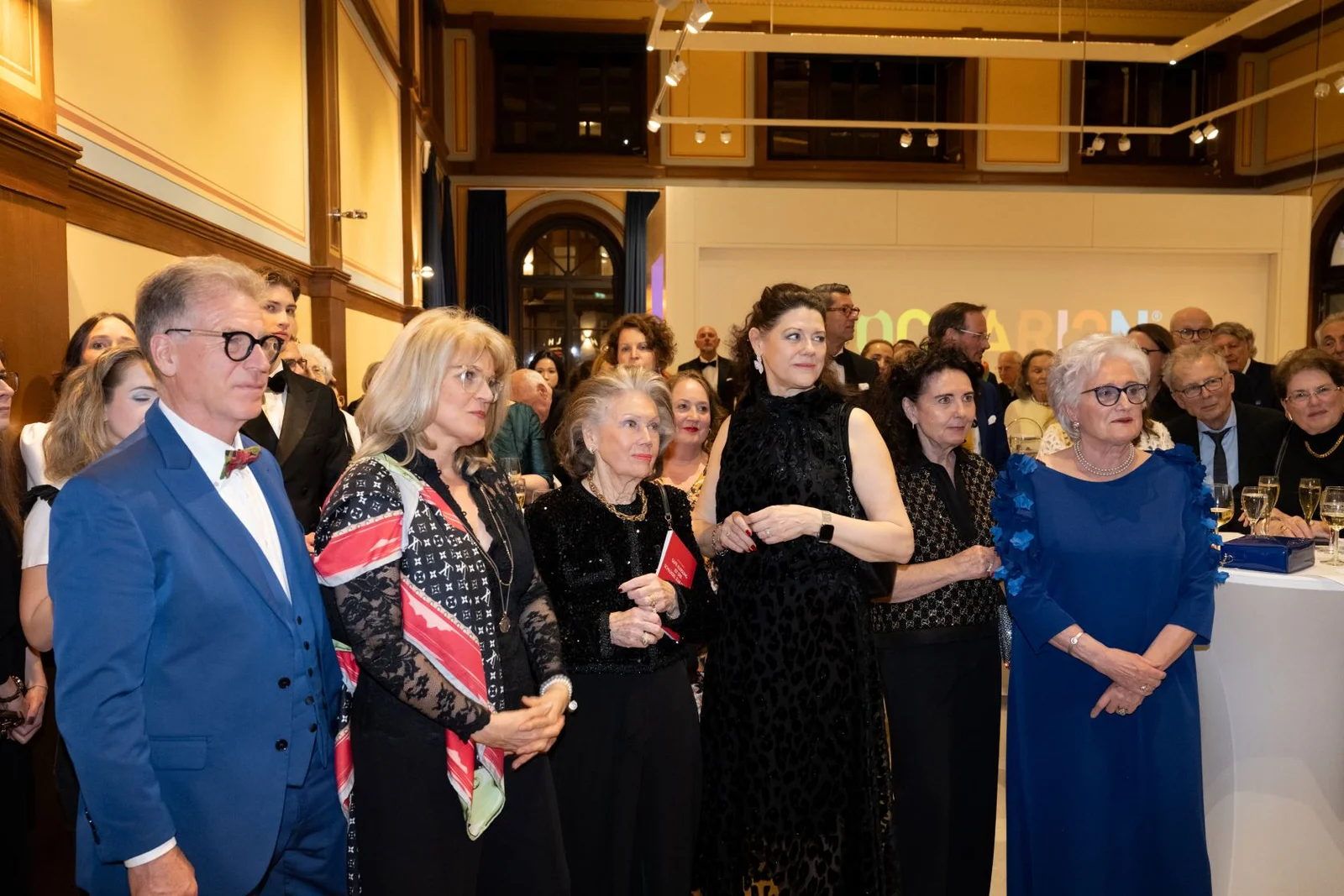 Menschen in eleganter Kleidung versammeln sich bei einer formellen Veranstaltung in einem stilvollen Raum mit hohen Decken und Kunstwerken.