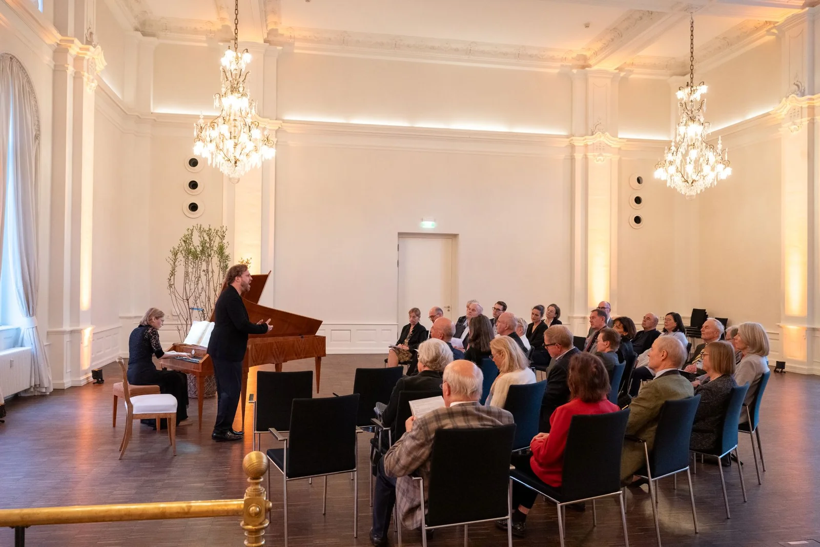 Ein eleganter Konzertsaal mit Zuhörern, die einem Klavierduo zuhören. Der Raum ist mit Kronleuchtern und großen Fenstern geschmückt.
