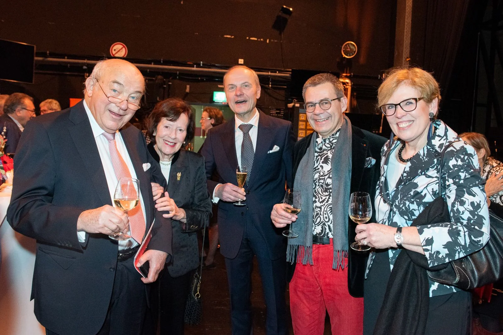 Eine Gruppe eleganter Menschen steht in einem Veranstaltungsraum. Sie lächeln und halten Weingläser in den Händen.