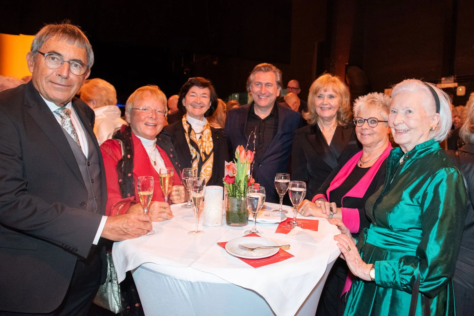 Eine Gruppe von Menschen steht um einen Tisch bei einer Veranstaltung. Sie stoßen mit Getränken an und lächeln in die Kamera.