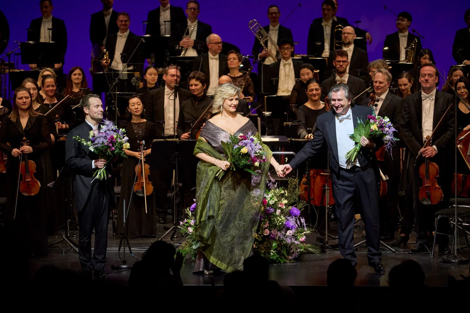 Eine Frau und zwei Männer nehmen nach einer Orchesteraufführung Blumensträuße entgegen. Musiker klatschen auf der Bühne.