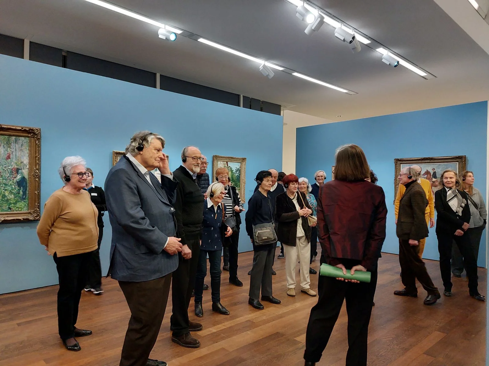 Der Freundeskreis des Festspielhauses Baden-Baden besucht eine Ausstellung im Museum Frieder Burda. Eine Führerin erklärt Gemälde an blau gestrichenen Wänden. Die Gruppe hört aufmerksam zu.
