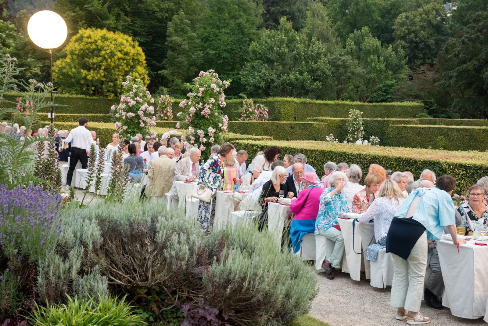 Der Freundeskreis des Festspielhauses Baden-Baden sitzt an langen Tischen im Freien, umgeben von grünen Gärten und blühenden Sträuchern, bei einer eleganten Abendveranstaltung.