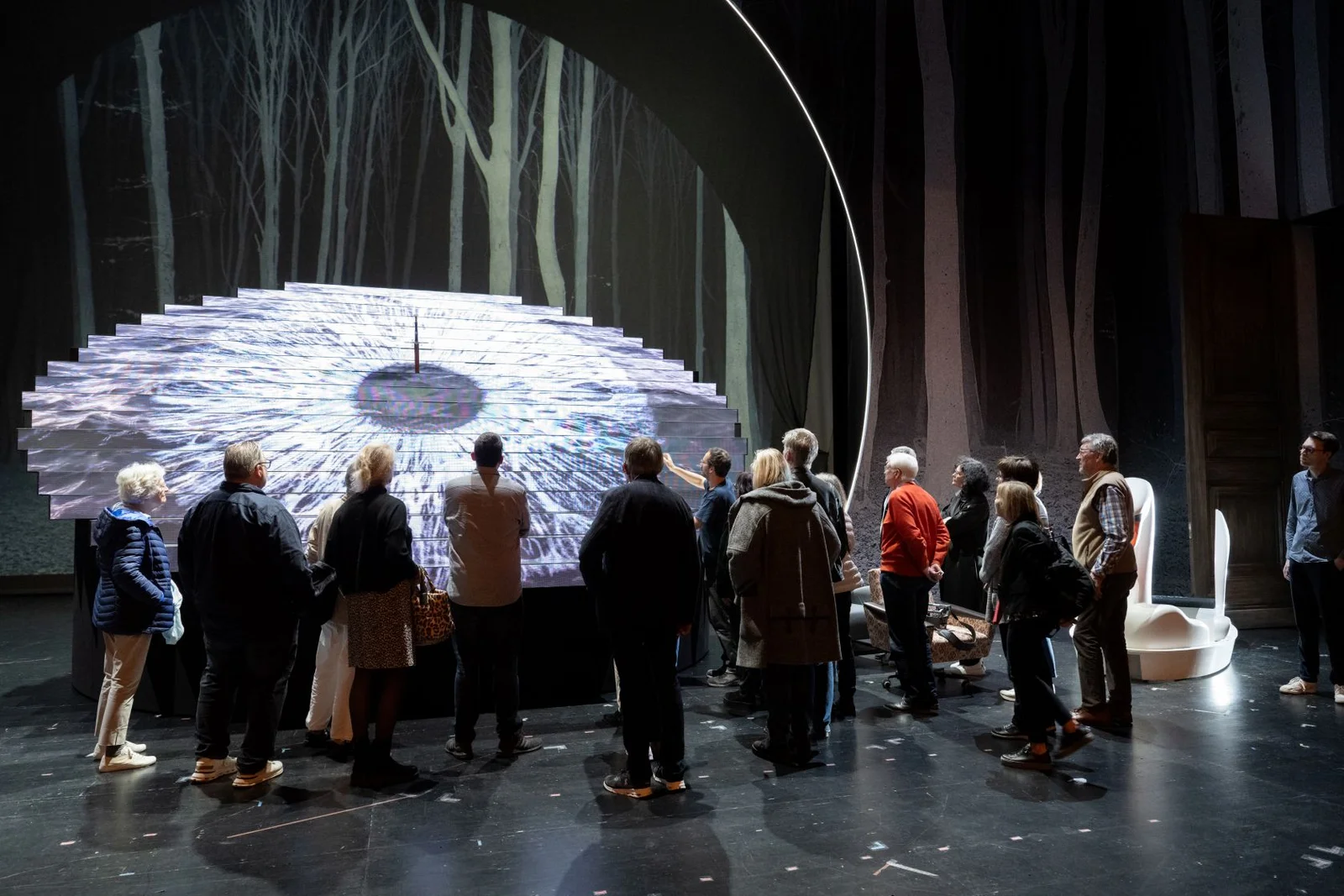 Der Freundeskreis des Festspielhauses Baden-Baden steht beim Probenbesuch der Oper "Lohengrin" auf einer Bühne. Sie versammeln sich um eine große, kreisförmige Treppeninstallation, die schwach beleuchtet ist.