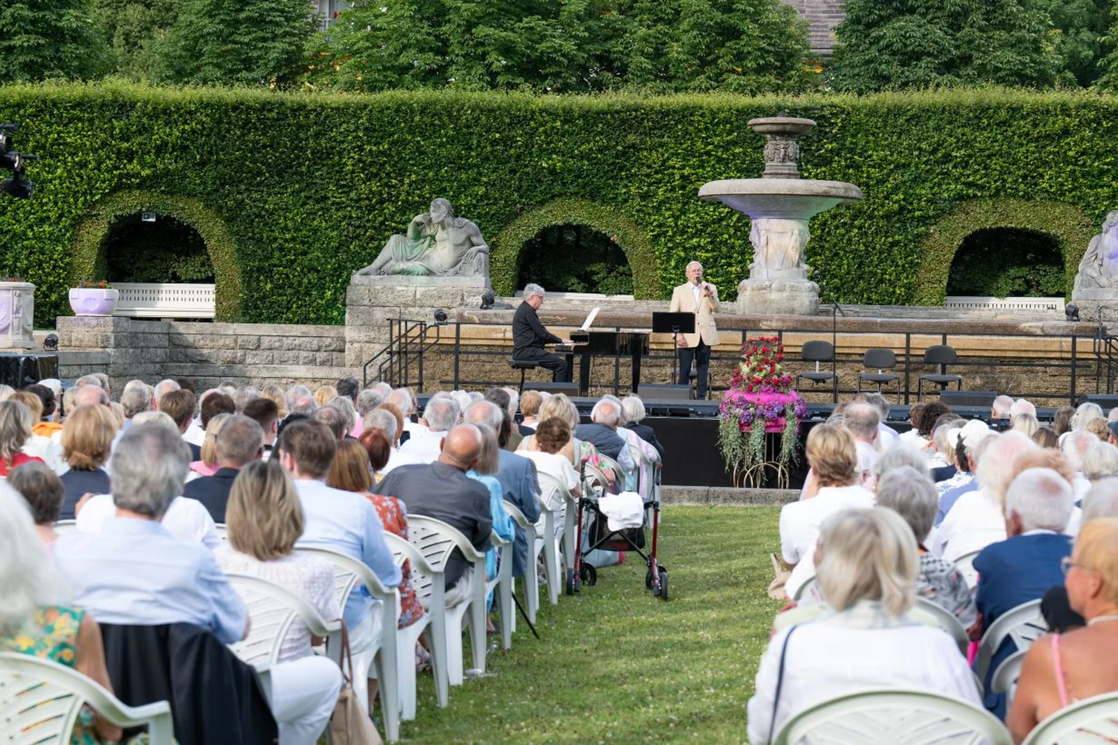 Mitglieder des Freundeskreises erleben beim Sommerfest ein Open-Air-Konzert. Ein Pianist und ein Sänger treten auf. Die Zuschauer sitzen auf Stühlen vor einer Hecke und einem Brunnen.