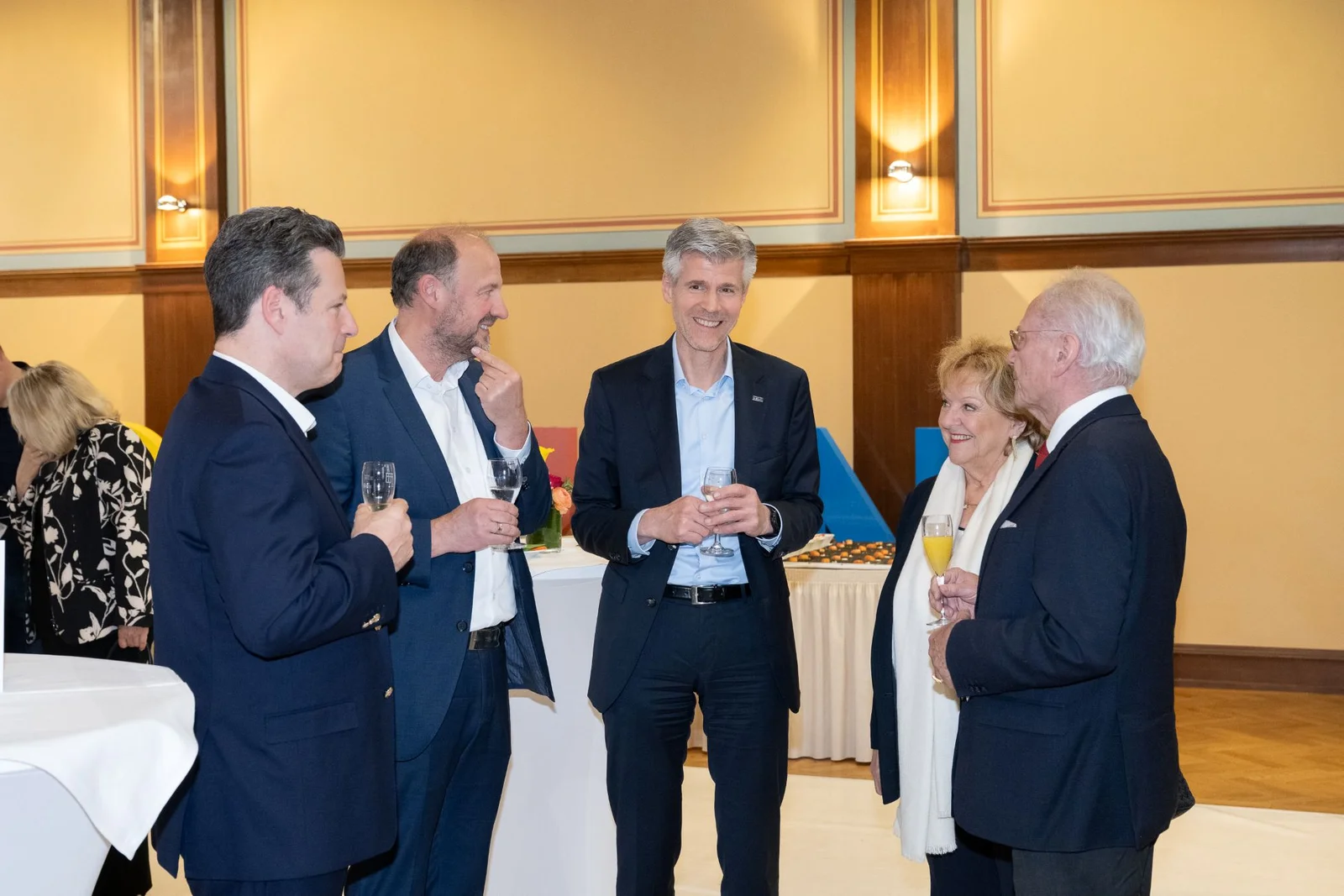 Fünf elegant gekleidete Personen stehen in einem Raum im Toccarion und unterhalten sich. Einige halten Sektgläser in der Hand.