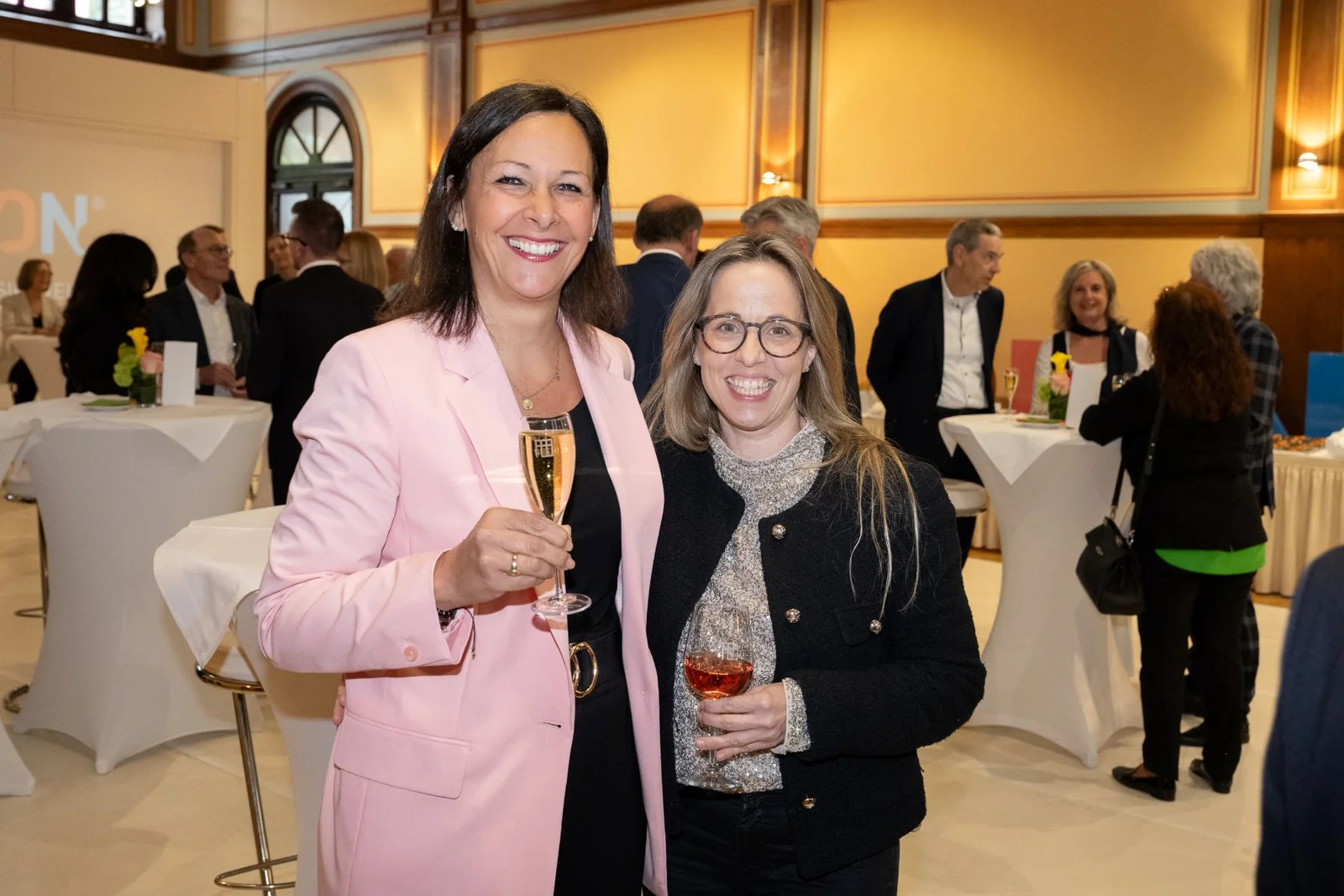 Zwei Personen lächeln und halten Getränke in einem Raum mit mehreren Menschen bei einem gesellschaftlichen Event. Elegante Kleidung und freundliche Atmosphäre.