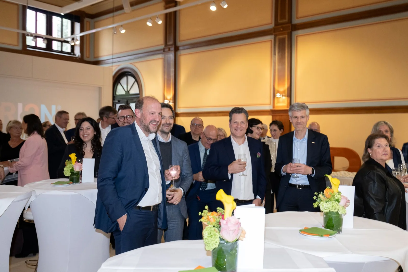 Eine Gruppe von Menschen in formeller Kleidung versammelt sich bei einem gesellschaftlichen Event in einem elegant dekorierten Raum mit Stehtischen.
