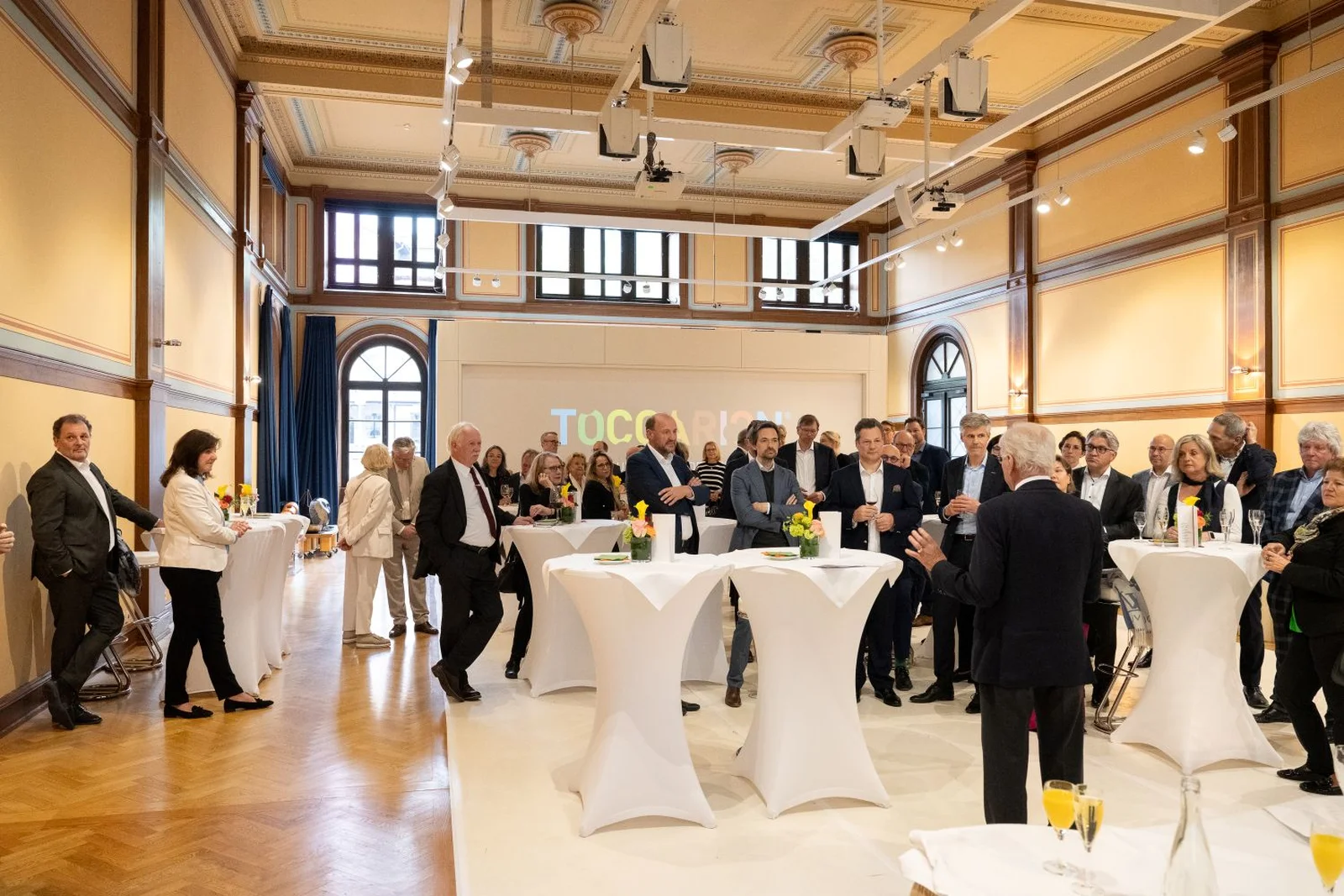 Menschen stehen an Stehtischen in einem großen Raum im Toccarion und unterhalten sich.
