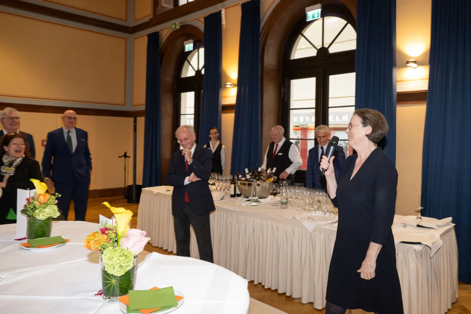 Geschäftsführerin Dr. Ursula Koners spricht in ein Mikrofon. Gäste stehen um sie herum und hören zu.
