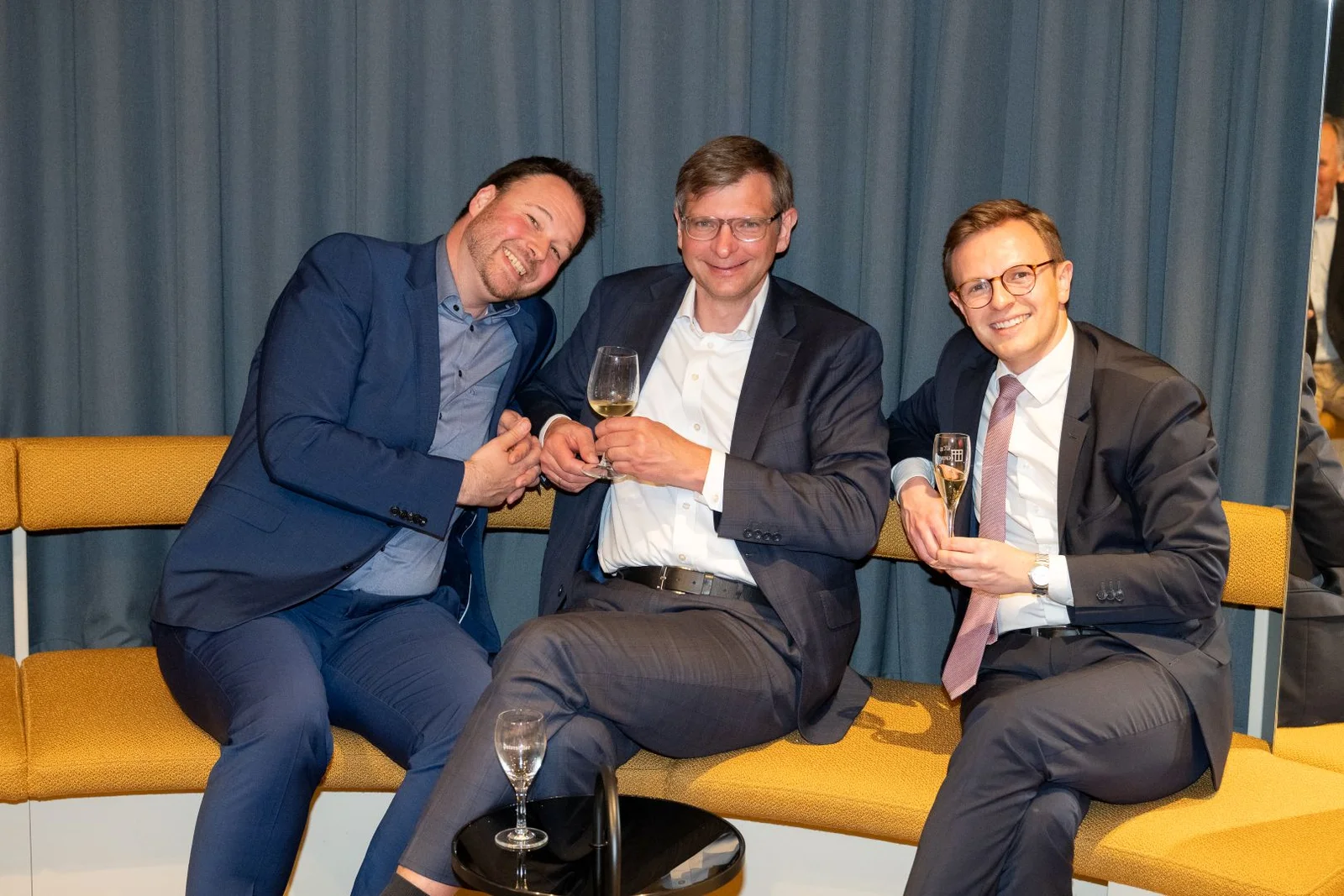 Drei Männer in Anzügen sitzen entspannt auf einer Bank und halten Sektgläser in der Hand. Sie lachen und wirken fröhlich.